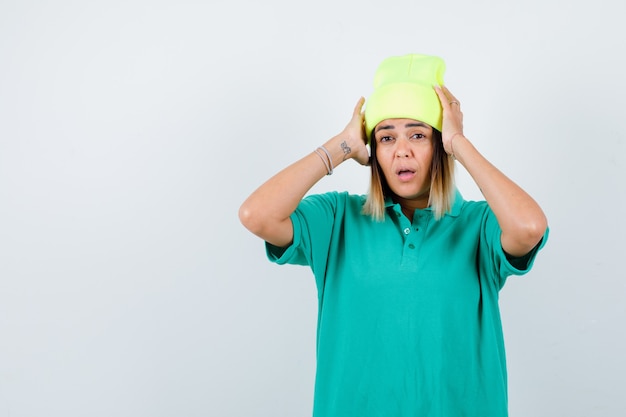 Portret van mooie vrouw met handen op het hoofd in polo t-shirt, muts en verbijsterd vooraanzicht