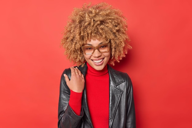 Portret van mooie vrouw met krullend borstelig haar draagt comfortabele coltrui leren jas houdt hand op schouder staat positief binnenshuis tegen felrode achtergrond drukt positieve emoties uit