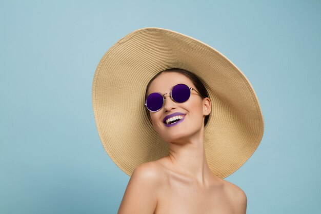 Portret van mooie vrouw met lichte make-up, hoed en zonnebril op blauwe studio