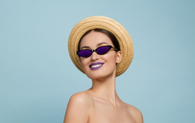 Portret van mooie vrouw met lichte make-up, hoed en zonnebril op blauwe studioachtergrond. Stijlvol en modieus merk en kapsel. Kleuren van de zomer. Schoonheid, mode, advertentieconcept. Kijkt naar de zijkant.