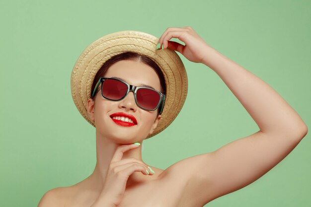 Portret van mooie vrouw met lichte make-up, hoed en zonnebril op groene ruimte. Stijlvol en modieus merk en kapsel. Kleuren van de zomer