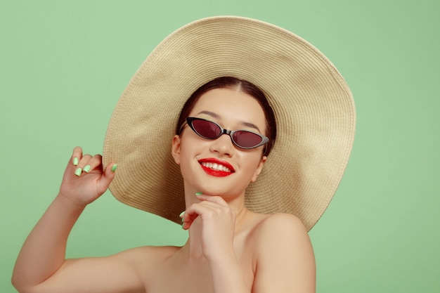 Portret van mooie vrouw met lichte make-up, hoed en zonnebril op groene studioachtergrond. stijlvol en modieus merk en kapsel. kleuren van de zomer. schoonheid, mode en advertentieconcept. lachend.