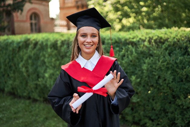 Portret van mooie vrouwelijke afgestudeerde in afstuderen gowm met diploma camera kijken.