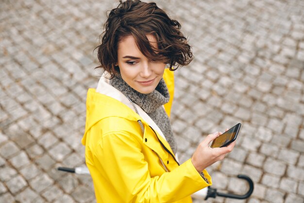 Portret van mooie vrouwelijke jaren '20 die op straatstenen met mobiele telefoon en paraplu in handen lopen die naar juiste route zoeken