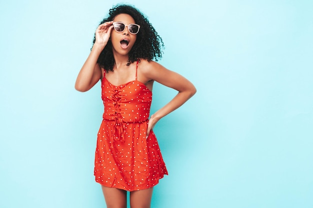 Portret van mooie zwarte vrouw met afro krullen kapsel glimlachend model gekleed in rode zomerjurk Sexy zorgeloze vrouw poseren in de buurt van blauwe muur in studio gelooid en vrolijk In zonnebril