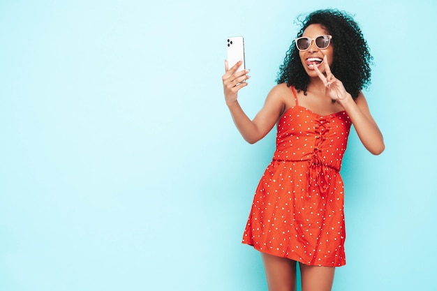 Portret van mooie zwarte vrouw met afro krullen kapsel glimlachend model gekleed in rode zomerjurk Sexy zorgeloze vrouw poseren in de buurt van blauwe muur in studio Gelooid en vrolijk Selfie nemen