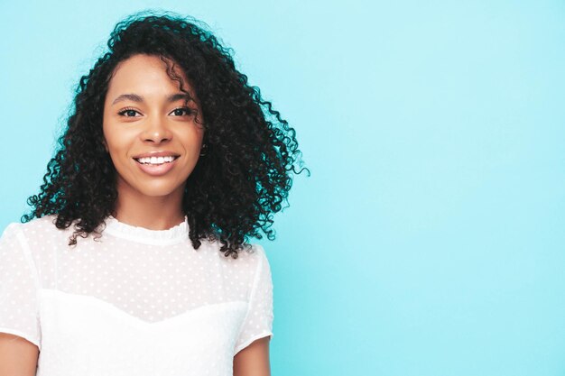 Portret van mooie zwarte vrouw met afro krullen kapsel glimlachend model gekleed in witte zomerjurk Sexy zorgeloze vrouw poseren in de buurt van blauwe muur in studio gelooid en vrolijk geïsoleerd