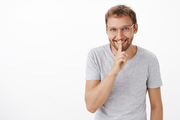 Portret van mysterieuze knappe man met borstelharen in glazen en grijs t-shirt glimlachend met enthousiasme zeggen shh, stil gebaar met wijsvinger over mond tonen