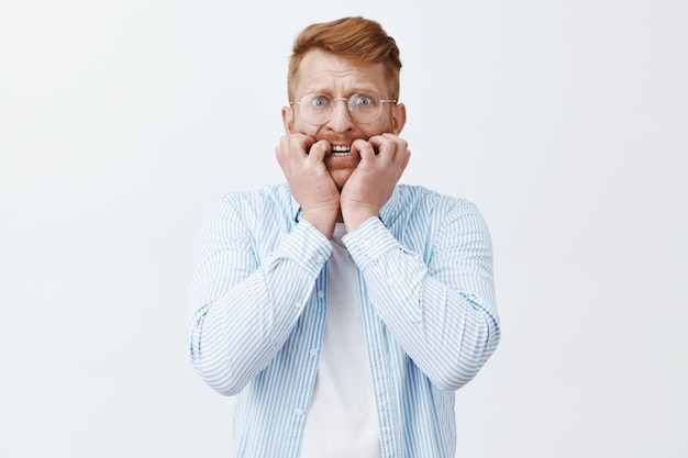 Portret van nerveuze boos roodharige volwassen man met borstelharen, vingernagels bijten, bukken en fronsen, onrustig en erg bezorgd over grijze muur