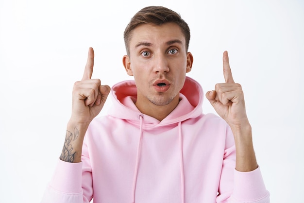 Gratis foto portret van nieuwsgierige knappe blonde man in roze hoodie wijzende vingers omhoog staren camera open mond geamuseerd ontdekte geweldige promo die vraag stelt geïnteresseerd en geïntrigeerd witte achtergrond