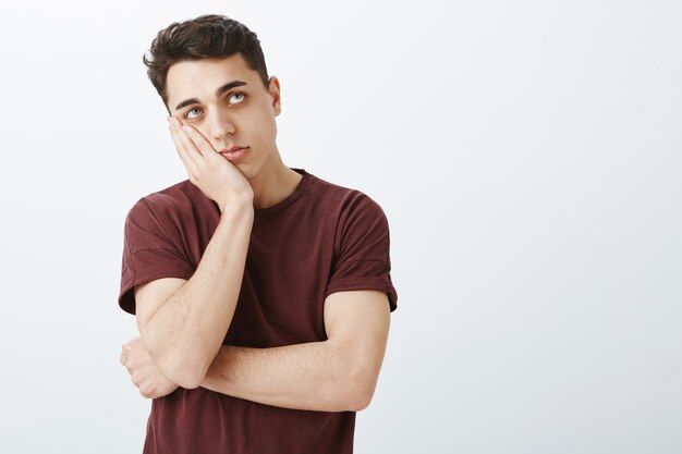 Portret van ongeduldige verveelde aantrekkelijke kerel in gewone rode t-shirt, leunend gezicht op handpalm en opzoeken