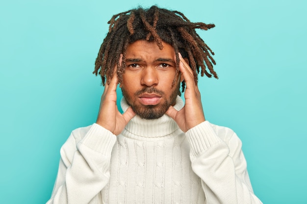 Portret van ongelukkige donkere man met dreadlocks, lijdt aan ondraaglijke headahce, houdt de handen op de slapen, draagt witte trui met kraag, geïsoleerd op blauwe achtergrond, vraagt om pijnstillers