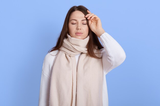 Portret van ongezonde vrouw gewikkeld in warme witte sjaal, haar hoofd aanraken, hoofdpijn, koorts en griepsymptomen lijden, ogen gesloten houden, geïsoleerd op lila muur.