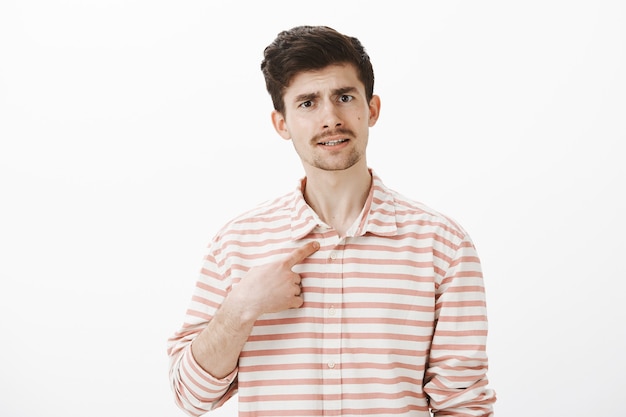 Portret van ontevreden aantrekkelijke man met snor, wijzend naar zichzelf met wijsvinger, wenkbrauwen optillen van verbazing, ruzie, beschuldigd van slechte dingen doen, gefrustreerd over grijze muur