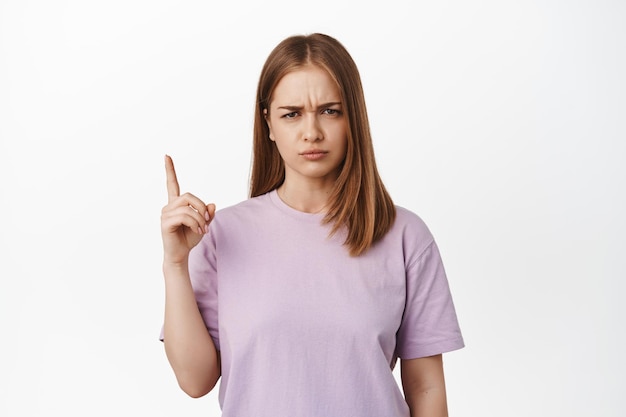 Portret van ontevreden blond tienermeisje, student klaagt, voelt zich oneerlijk, wijst met de vinger omhoog en fronst wenkbrauwen van streek, klagend over een slechte, witte achtergrond.