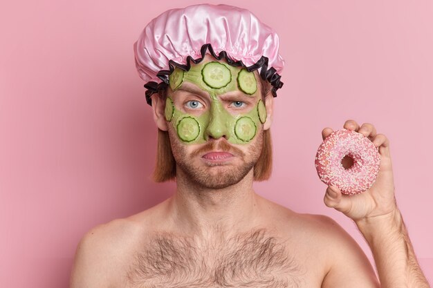 Portret van ontevreden nors man met snor fronst gezicht houdt heerlijke donut staat topless binnen past gezichts groen masker komkommer plakjes voor huidverjonging.