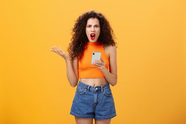 Portret van ontevreden pissige en geïrriteerde blanke vrouw met krullend kapsel in rode lippenstift en oranje bijgesneden top met smartphone en gebarend met palm geïrriteerd klagend over stom bericht.