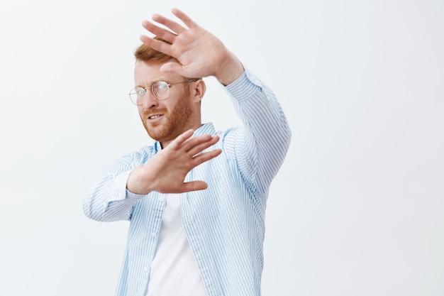 Portret van ontevreden, verontruste en bezorgde beroemde mannelijke ondernemer met rood haar, die zich afwendt, gezicht bedekt met opgeheven handpalmen, probeert zich te verbergen voor paparazzi over grijze muur