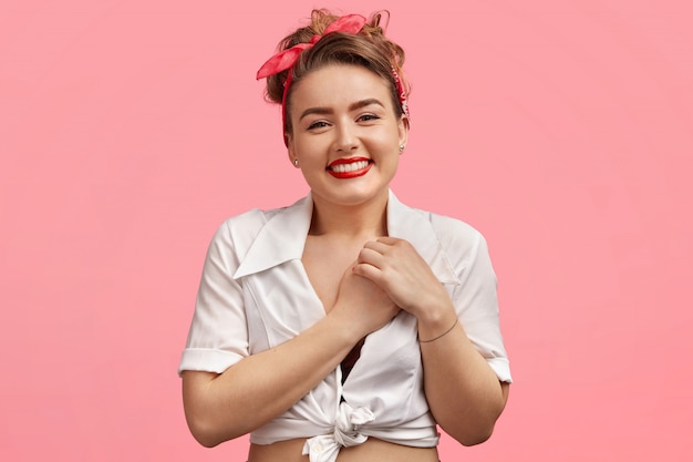 Portret van opgetogen mooie vrouw met make-up, rode lippen, houdt de handen op het hart, lacht vrolijk naar de camera, vormt tegen roze