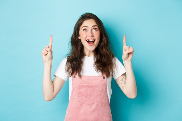 Portret van opgewonden brunette meisje in zomeroutfit, kijkend en wijzende vingers omhoog met blij gezicht, promo-aanbieding bekijken, coole advertentie tonen, blauwe achtergrond