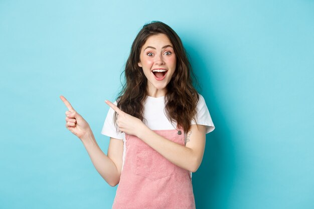 Portret van opgewonden gelukkige vrouw met donkere glanzende krullen, gefascineerd glimlachend, wijzende vingers naar links naar logobanner, demonstreren een advertentie, staande over blauwe achtergrond.