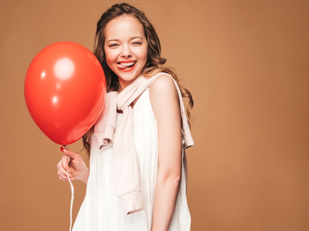 Portret van opgewonden jonge meisje poseren in trendy zomer witte jurk. Glimlachende vrouw met het rode ballon stellen. Model klaar voor feest