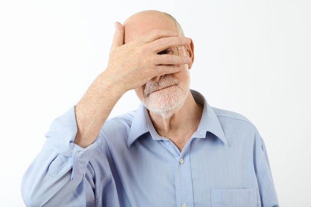 Portret van oudere gepensioneerde blanke man in blauw shirt met hand op zijn gezicht, ogen bedekt en gluren door gespleten vingers, beschaamd. Menselijke gezichtsuitdrukkingen en lichaamstaal