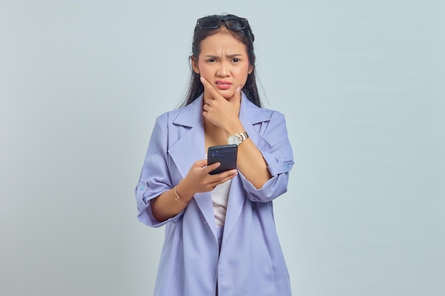Portret van peinzende jonge aziatische vrouw die mobiele telefoon gebruikt die op witte achtergrond wordt geïsoleerd