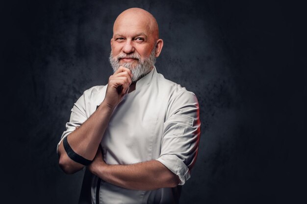 Portret van peinzende oude man chef-kok gekleed in uniform poseren tegen een donkere achtergrond.