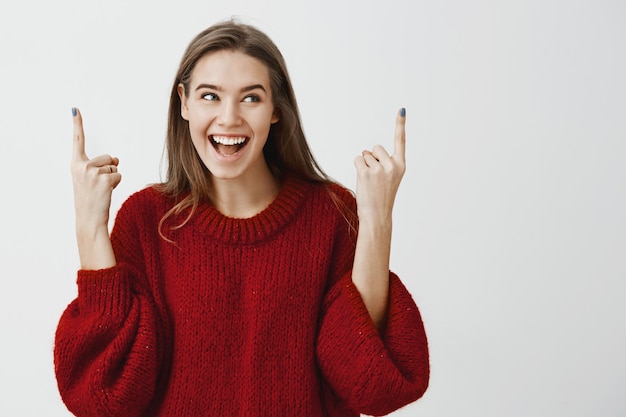 Portret van positieve vrolijke charmante vrouw in rode losse trui, wijsvingers opheffen en omhoog wijzen, opzij kijken met positieve gelukkige glimlach, poseren voor reclame en vertellen over goed item