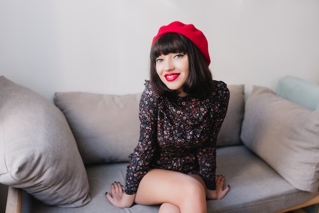 Portret van prachtig meisje met kort donker haar, zittend op de grijze bank en verleidelijk glimlachen. Aantrekkelijke jonge lachende vrouw met trendy kapsel Franse outfit dragen graag poseren in lichte kamer