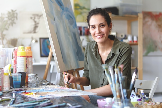 Portret van prachtige opgewonden jonge brunette vrouwelijke kunstenaar in casual blouse van kaki kleur, het mengen van olieverf op palet met schildermes, gepassioneerd over haar beroep en creatieproces