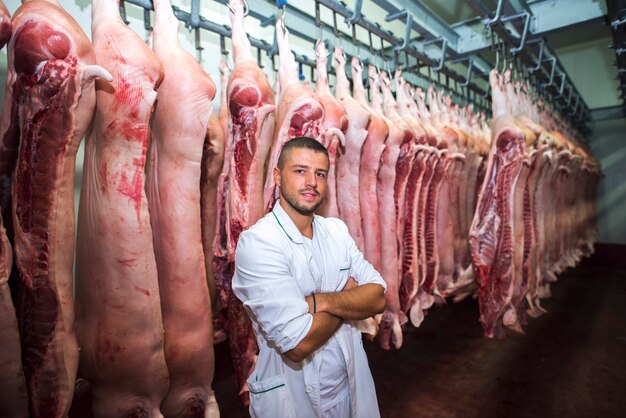 Portret van professionele slager in koude opslag van de fabriek met armen gekruist met varkenskarkas achter