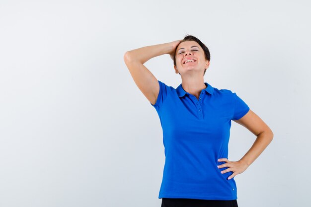 Portret van rijpe vrouw die hand op hoofd in blauw t-shirt houdt en gelukkig vooraanzicht kijkt