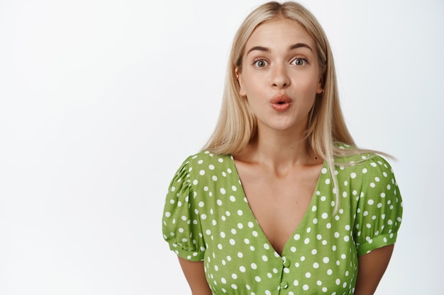 Portret van schattig blond meisje ziet er nieuwsgierig uit, tuit lippen en kijkt geïntrigeerd, staande in groene jurk op wit.