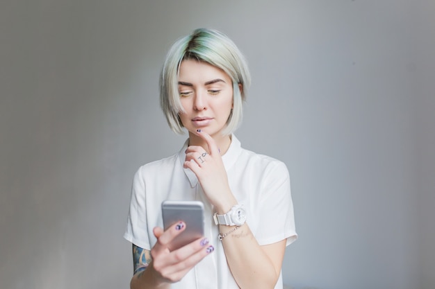 Portret van schattig meisje met grijze korte haarstijl staande op een grijze achtergrond. Ze draagt een witte jurk en lichte make-up, houdt de telefoon vast.