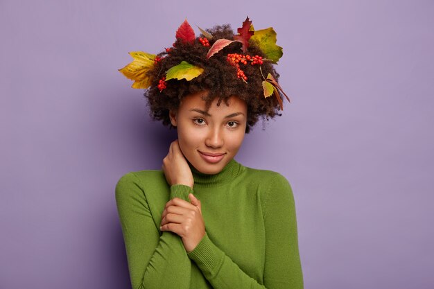 Portret van schattige Afro-Amerikaanse dame raakt nek, kijkt rustig naar de camera, draagt casual poloneck, herfstbladeren, bessen in krullend haar voor een perfect kapsel tevreden