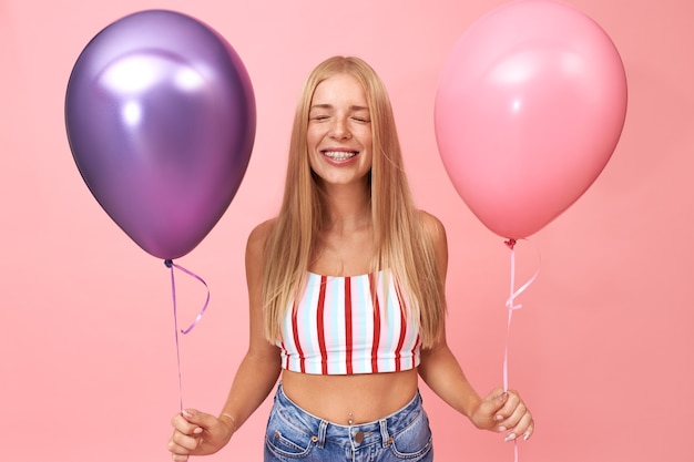 Portret van schattige gelukkige jonge Europese vrouw bretels en crop top sluiten ogen dragen tijdens het maken van wens op haar verjaardag, genieten van feest met vrienden, glimlachend