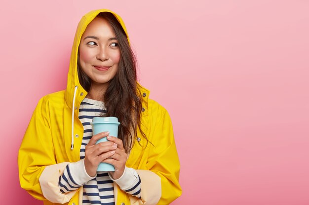 Portret van schattige jonge vrouw draagt regenjas, verwarmt met warme drank, kijkt gelukkig opzij, heeft kuiltjes op rouge wangen