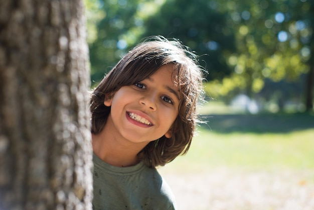 Portret van schattige kleine jongen in het park. Donkerharige lachende jongen die uit de boom gluurt en naar de camera kijkt. Familie, liefde, jeugdconcept
