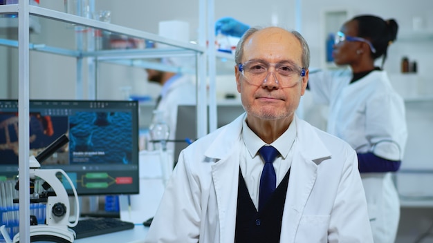 Portret van senior ervaren wetenschapper man die lacht op camera in modern uitgerust lab. multi-etnisch team dat virusevolutie onderzoekt met behulp van hightech- en scheikundige hulpmiddelen voor wetenschappelijk onderzoek, vaccin d