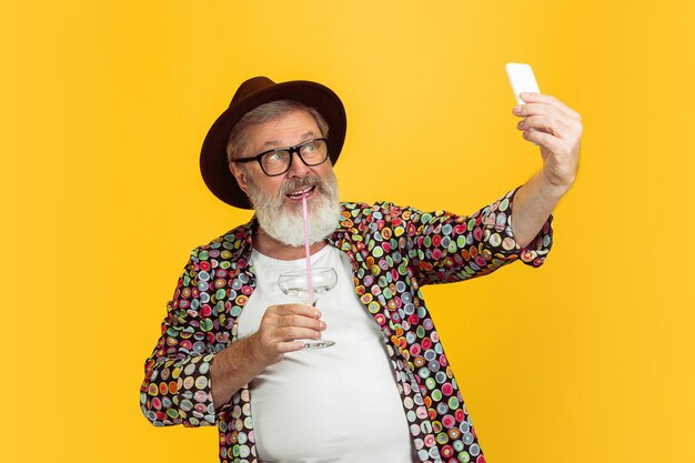 Portret van senior hipster man met behulp van apparaten, gadgets geïsoleerd op gele studio achtergrond. Tech en vreugdevolle ouderen levensstijl concept.