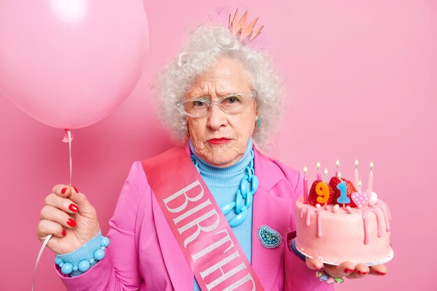 Portret van serieuze grootmoeder viert 91e verjaardag met heerlijke taart met brandende kaarsen opgeblazen ballon gekleed in feestelijke outfit ziet er helaas uit als oud