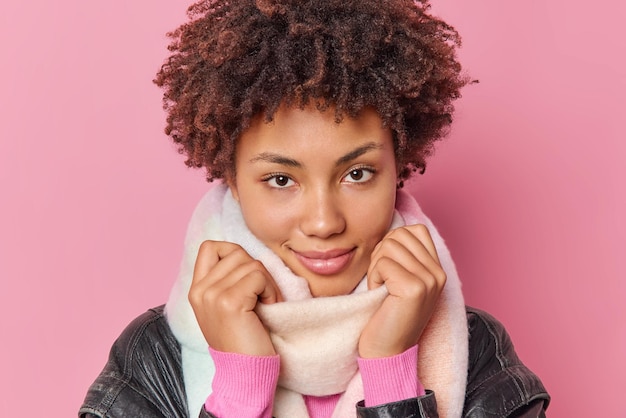 Portret van serieuze mooie vrouw met krullend borstelig haar draagt warme sjaal om nek tijdens koude winterdag kijkt direct naar camera geïsoleerd over roze achtergrond. Mensen en kleding concept.