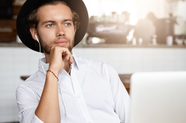 Portret van serieuze student met wit overhemd en zwarte hoed met een doordachte uitdrukking, vooruitkijkend terwijl hij luistert naar audioboek op oortelefoons, binnenshuis zit voor open laptop pc