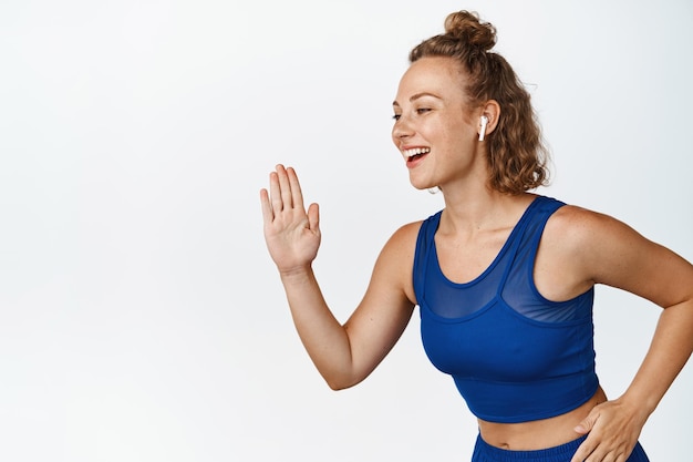 Portret van sportvrouw die rent en muziek luistert in draadloze oortelefoons. Vrouwelijke atleet joggen en lachen, het dragen van activewear, witte achtergrond.