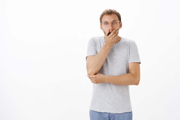 Portret van sprakeloos bezorgd en schudde ongeschoren man in glazen die geopende mond bedekten met bezorgd handpalm staren