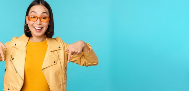 Portret van stijlvol aziatisch meisje draagt een zonnebril, glimlacht en wijst met de vingers naar beneden, laat advertenties zien die op een blauwe achtergrond staan