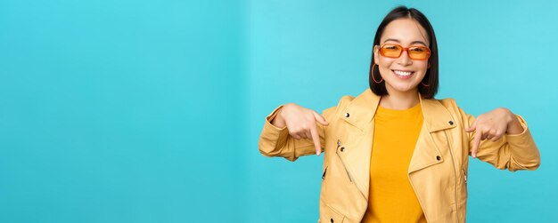 Portret van stijlvol aziatisch meisje draagt een zonnebril, glimlacht en wijst met de vingers naar beneden, laat advertenties zien die op een blauwe achtergrond staan