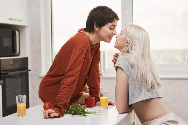 Portret van stijlvolle en aantrekkelijke lesbienne in rode pullover zitten aan de keukentafel terwijl kussen vriendin en gelukkig lachend tijdens het ontbijt. Meisje vond eindelijk haar zielsverwant